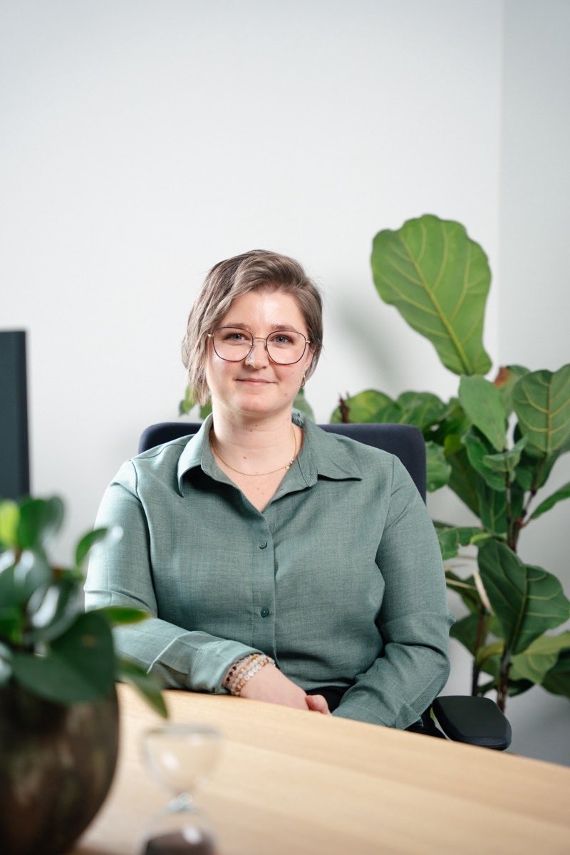 Mitarbeiterin Nelli Giesbrecht sitzt lächelnd am Schreibtisch mit großer Zimmerpflanze im Hintergrund